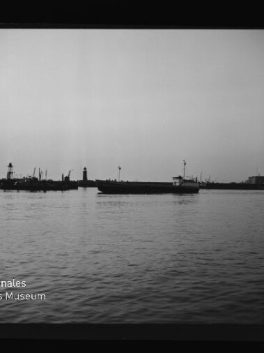 Schwarzweißaufnahme eines Frachtschiffs vor einer Hafenmole mit Leuchtturm und Kranen, Museumsschriftzug unten links.