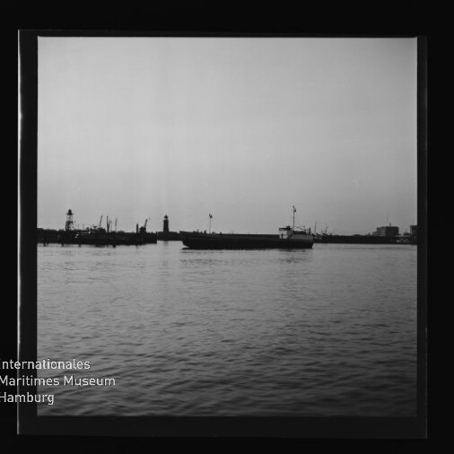 Schwarzweißaufnahme eines Frachtschiffs vor einer Hafenmole mit Leuchtturm und Kranen, Museumsschriftzug unten links.