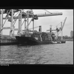 Containerschiff am Kai unter großen Containerkränen, Schlepper im Vordergrund; Schwarzweißfoto mit Wasserzeichen Internationales Maritimes Museum Hamburg