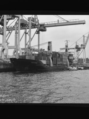 Containerschiff am Kai unter großen Containerkränen, Schlepper im Vordergrund; Schwarzweißfoto mit Wasserzeichen Internationales Maritimes Museum Hamburg