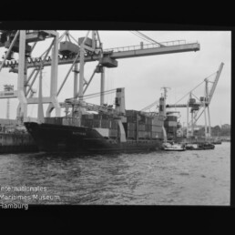 Containerschiff am Kai unter hohen Hafenkränen, beladene Containerstapel und ein Schlepper im Vordergrund.