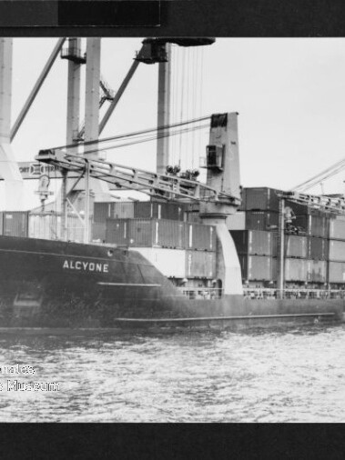 Containerschiff ALCYONE am Hafen mit großen Verladekränen, gestapelten Containern und einem kleinen Schlepper.