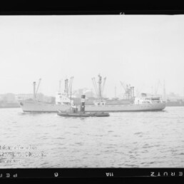 Frachtschiff, das von einem kleinen Schlepper begleitet wird, vor Hafenpier mit Kränen und Industrieanlagen im Hintergrund.