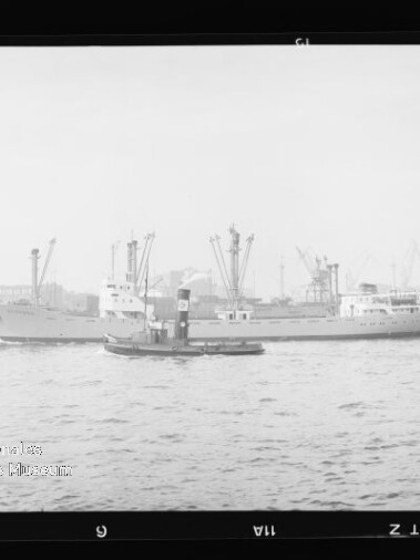 Frachtschiff, das von einem kleinen Schlepper begleitet wird, vor Hafenpier mit Kränen und Industrieanlagen im Hintergrund.