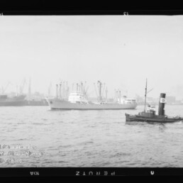 Frachtschiff in der Hafeneinfahrt, im Vordergrund ein Schlepper und im Hintergrund Hafenkräne sowie weitere Schiffe.