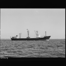 Schwarzweißfoto eines Frachtschiffs mit Ladekränen auf ruhiger See; unten links Logo des Maritimen Museums Hamburg.