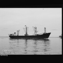 Schwarzweiß-Seitenansicht eines Frachtschiffs mit mehreren Ladekranen auf ruhigem Wasser; Wasserzeichen: Internationales Maritimes Museum Hamburg.