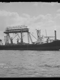 Frachtschiff im Hafen vor einer großen Krananlage mit sichtbarem Schriftzug, ruhiges Wasser und wolkiger Himmel.