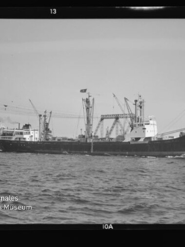 Frachtschiff fährt am Hafen vorbei, im Hintergrund Kräne und Werft, Wasser im Vordergrund und Museumssignet unten links.