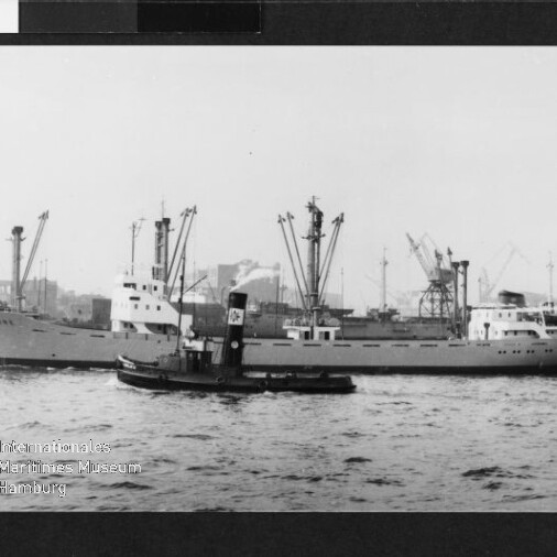 Graustufenfoto eines Frachtschiffs mit mehreren Ladekränen und einem kleinen Schlepper im Vordergrund vor Hafenkränen.
