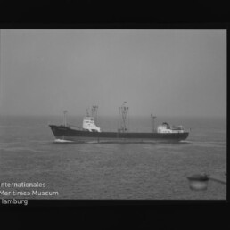 Schwarzweißaufnahme eines Frachtschiffs mit Ladekränen auf ruhiger See, Filmrand und Museumslogo links unten.