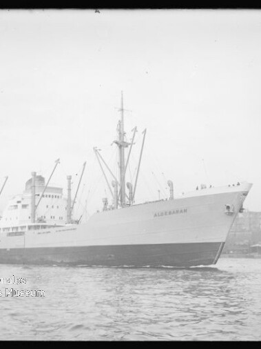 Großes Frachtschiff mit Ladekränen und sichtbarem Schriftzug am Bug fährt in einen Hafen, im Hintergrund Industrieanlagen.