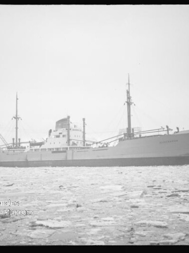 Frachtschiff in Seitenansicht auf zugefrorenem Wasser vor Hafenanlagen, kleines Museum-Logo links unten.