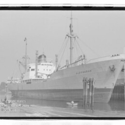 Frachtschiff ALDEBARAN liegt am Kai, sichtbarer Bug mit Pollern, kleines Beiboot im Wasser; Schwarzweißaufnahme mit Museumslogo.