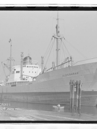 Frachtschiff ALDEBARAN liegt am Kai, sichtbarer Bug mit Pollern, kleines Beiboot im Wasser; Schwarzweißaufnahme mit Museumslogo.