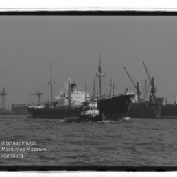 Schlepper begleitet großes Frachtschiff vor Hafenkränen, mit Schriftzug Internationales Maritimes Museum Hamburg unten links.