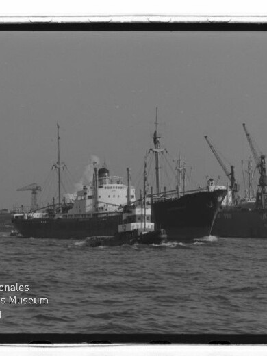 Schlepper begleitet großes Frachtschiff vor Hafenkränen, mit Schriftzug Internationales Maritimes Museum Hamburg unten links.