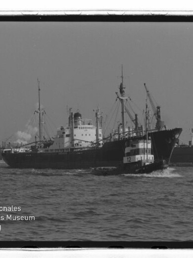 Frachtschiff im Hafen, davor ein Schlepper mit Nummer 12, Hafenkräne im Hintergrund und sichtbarer Museumsschriftzug.