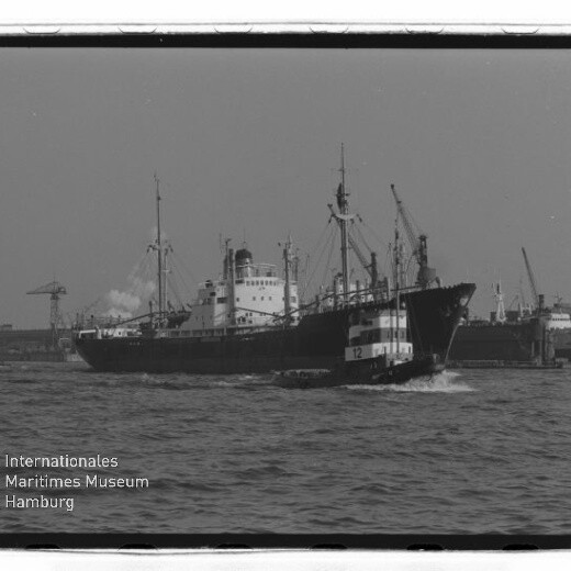 Frachtschiff im Hafen, davor ein Schlepper mit Nummer 12, Hafenkräne im Hintergrund und sichtbarer Museumsschriftzug.