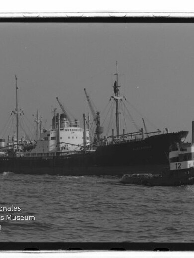 Großes Frachtschiff mit Ladekränen im Hafen, im Vordergrund ein Schlepper mit Nummer 12; Museumslogo unten links.