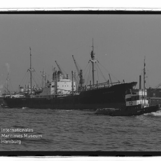 Großes Frachtschiff mit Ladekränen im Hafen, im Vordergrund ein Schlepper mit Nummer 12; Museumslogo unten links.
