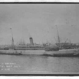 Passagierdampfschiff mit hohem Schornstein und Masten im Hafen, umgeben von kleinen Booten, historische Schwarzweißaufnahme.