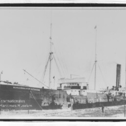 Frachtschiff mit Schornstein und Masten am Kai, Rumpf beschriftet und Wasserzeichen des Internationalen Maritimen Museums Hamburg.