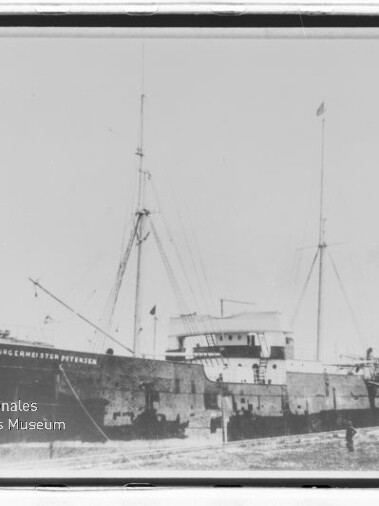 Frachtschiff mit Schornstein und Masten am Kai, Rumpf beschriftet und Wasserzeichen des Internationalen Maritimen Museums Hamburg.