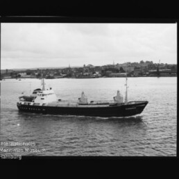 Schwarzweißbild eines Frachtschiffs namens Alexandria, das vor der Uferlinie einer Hafenstadt mit Kirchtürmen fährt.