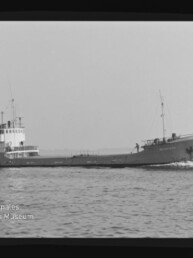 Kleines Frachtschiff in Seitenansicht fährt über ruhige See, mit Aufbauten achtern und Museum-Logo unten links.