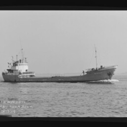 Kleines Frachtschiff in Seitenansicht fährt über ruhige See, mit Aufbauten achtern und Museum-Logo unten links.