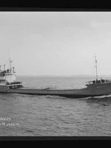 Kleines Frachtschiff mit Aufbauten achtern und Mast vorn auf ruhiger See; Schwarzweißfoto mit Wasserzeichen unten links.