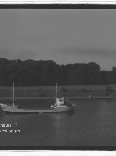 Kleines Frachtschiff mit zwei Masten auf ruhigem Wasser vor baumbewachsenem Ufer; Museumswasserzeichen unten links