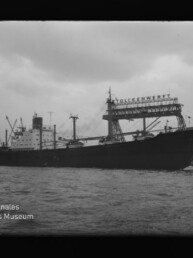 Großes Frachtschiff im Hafen vor Werftkränen, trüber Himmel; Museumslogo Internationales Maritimes Museum links unten.