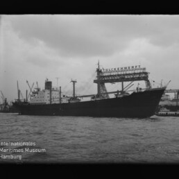 Großes Frachtschiff im Hafen vor Werftkränen, trüber Himmel; Museumslogo Internationales Maritimes Museum links unten.