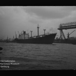 Frachtschiff im Hafen vor Kranen und Werftbauten bei bewölktem Himmel, ruhiges Wasser und Museumslogo unten links