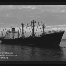Großes Frachtschiff mit zahlreichen Ladebäumen auf ruhigem Wasser, schwarz-weiß-Foto mit Museumslogo Internationales Maritimes Museum Hamburg.