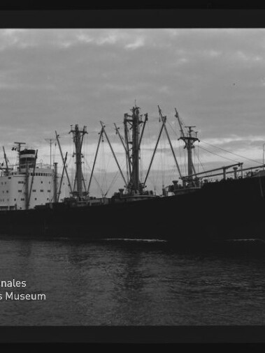 Großes Frachtschiff mit zahlreichen Ladebäumen auf ruhigem Wasser, schwarz-weiß-Foto mit Museumslogo Internationales Maritimes Museum Hamburg.