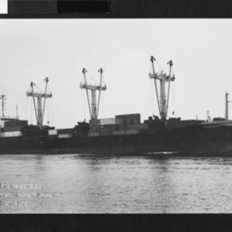 Schwarzweißfoto eines Frachtschiffs mit drei Ladekränen und Containern auf ruhigem Wasser, Museumsstempel unten links.