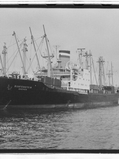 Großes Frachtschiff BUNTENSTEIN mit mehreren Ladekränen im Hafen und kleinem Lotsenboot am Heck.