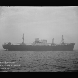 Schwarzweißfoto eines Frachtschiffs mit Aufbauten und Ladekränen auf ruhiger See, Museumswasserzeichen unten links.