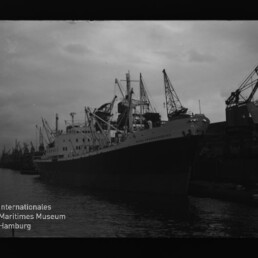 Großes Frachtschiff am Kai, mehrere Ladekräne im Hafen; Schriftzug Internationales Maritimes Museum Hamburg links unten.