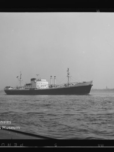 Frachtschiff in Schwarzweißaufnahme auf ruhiger See vor flachem Ufer, mit Museumswasserzeichen links unten.