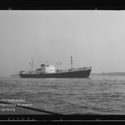 Frachtschiff in Schwarzweißaufnahme auf ruhiger See vor flachem Ufer, mit Museumswasserzeichen links unten.