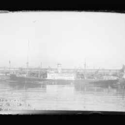 Schwarzweißaufnahme mehrerer Frachtschiffe mit Schornstein am Hafen vor Kränen und Lagerhäusern, Spiegelung im Wasser.