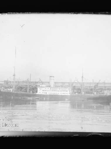 Schwarzweißaufnahme mehrerer Frachtschiffe mit Schornstein am Hafen vor Kränen und Lagerhäusern, Spiegelung im Wasser.