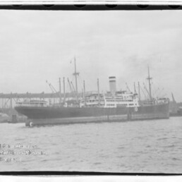 Großes Passagier- und Frachtschiff mit hohem Schornstein am Kai, Kräne und Hafenbrücke im Hintergrund.