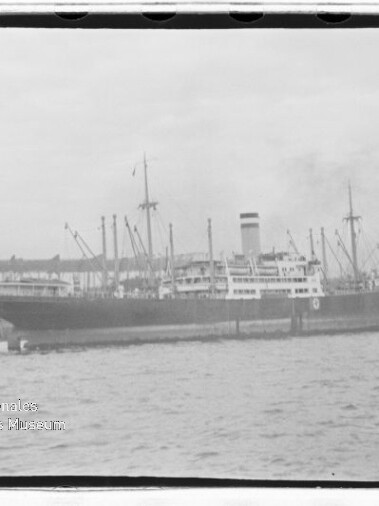 Großes Passagier- und Frachtschiff mit hohem Schornstein am Kai, Kräne und Hafenbrücke im Hintergrund.