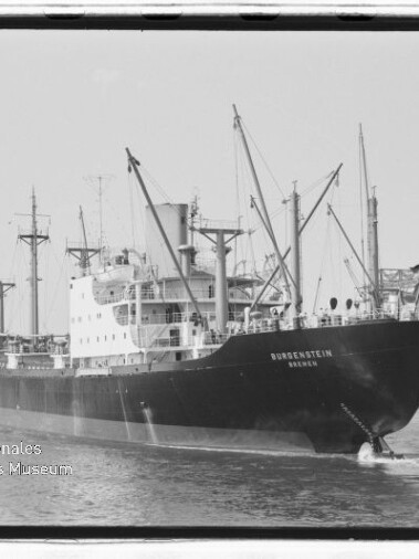 Schwarzweiß‑Frachter mit Ladekränen im Hafen, Name am Bug sichtbar und Museumswasserzeichen links unten.