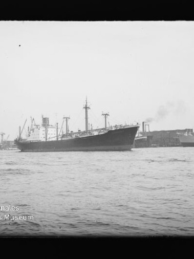 Frachtschiff mit Schornstein und Ladekränen im Hafen, Kaimauern und Kräne im Hintergrund; Wasserzeichen unten links.